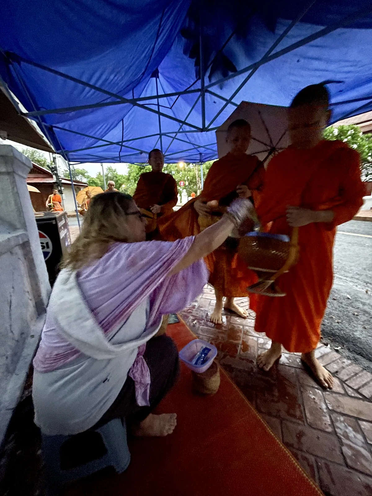 A woman in white kneeling and giving alms to barefoot monks in saffron robes with bowls during the Tak Bat