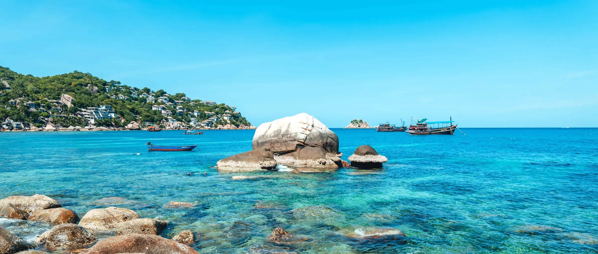 Shark Bay panorama, granite islet at the centre, dive boats anchored, the hill resorts of Koh Tao behind