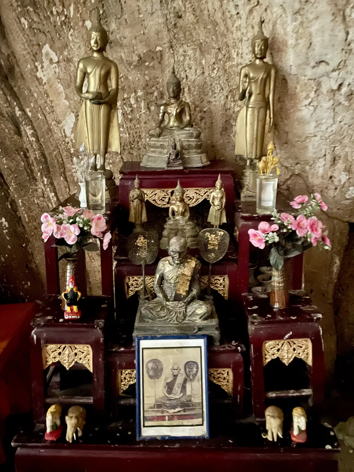 A detailed altar inside Phraya Nakhon Cave with golden Buddha statues, a seated monk figure and pink flowers